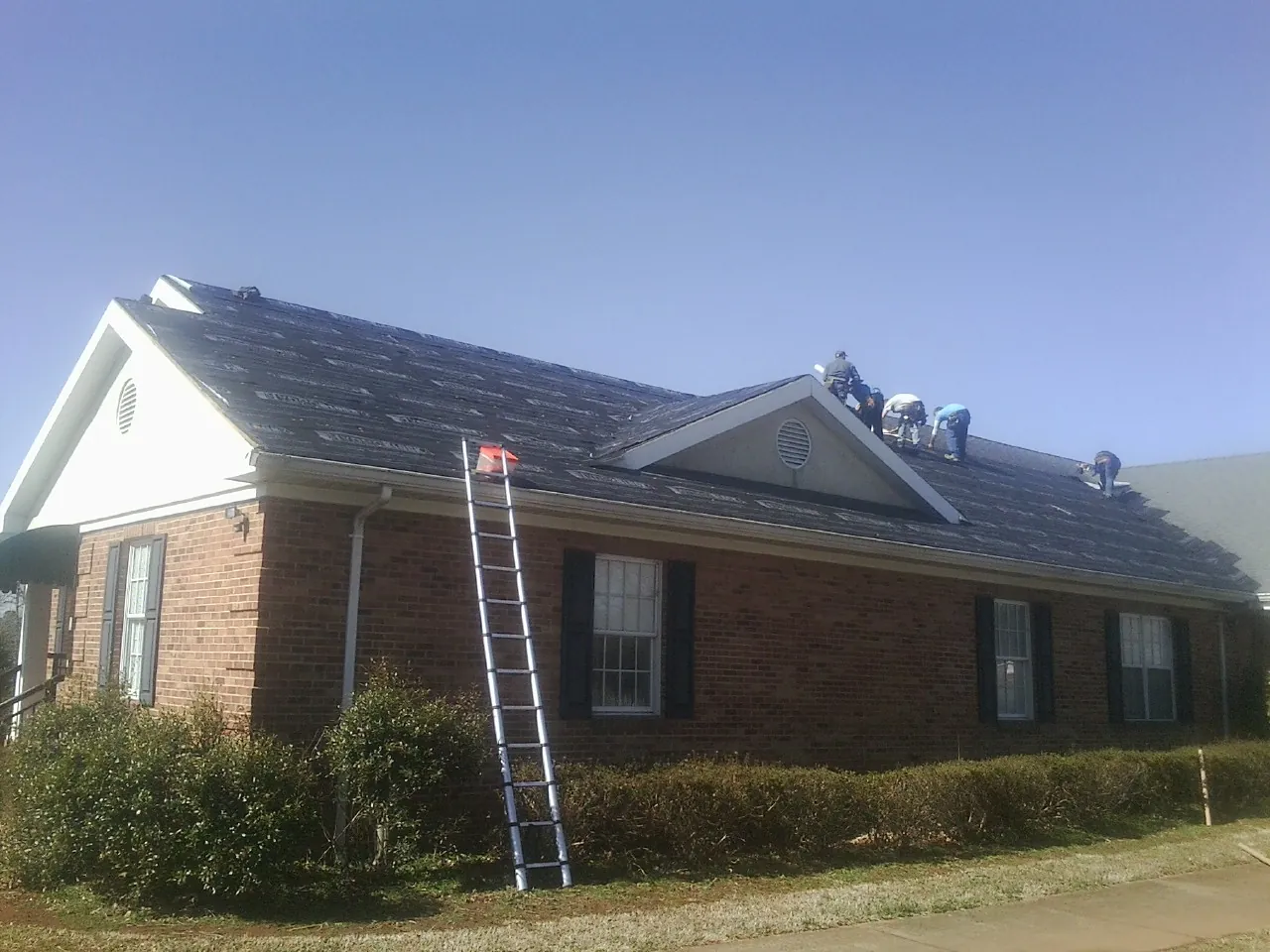 Crew tearing shingles off a brick ranch roof with a ladder set — Metal Roof Repair work by our crew in Farmington